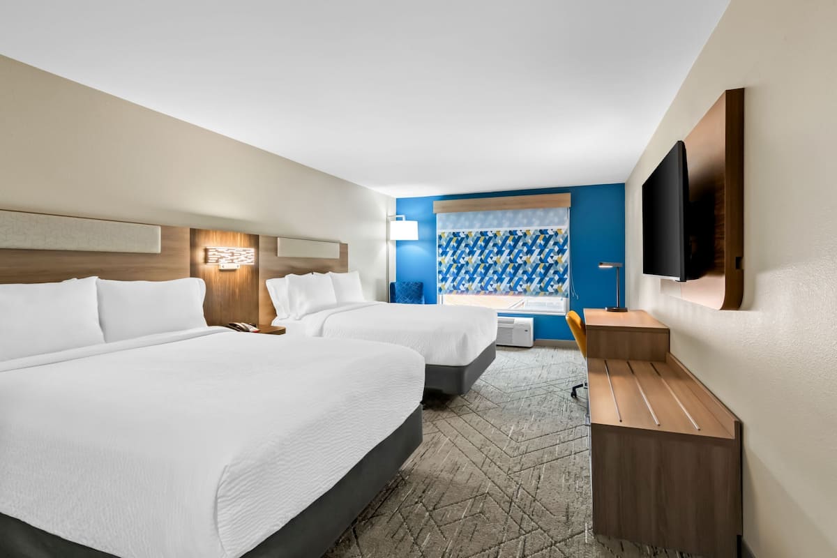A hotel room with two beds, a desk, a flat-screen TV, and a window with patterned curtains.