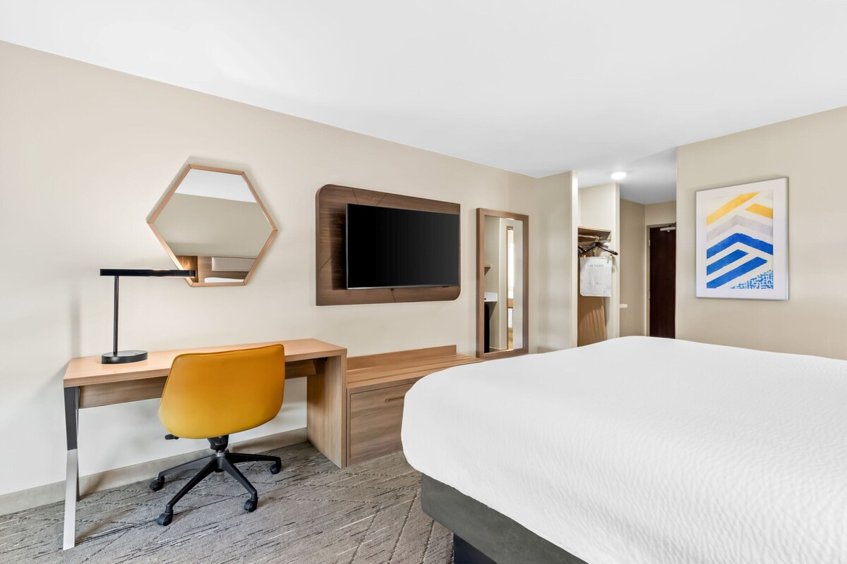 A hotel room with a bed, a desk with a chair, a mirror, and a television.
