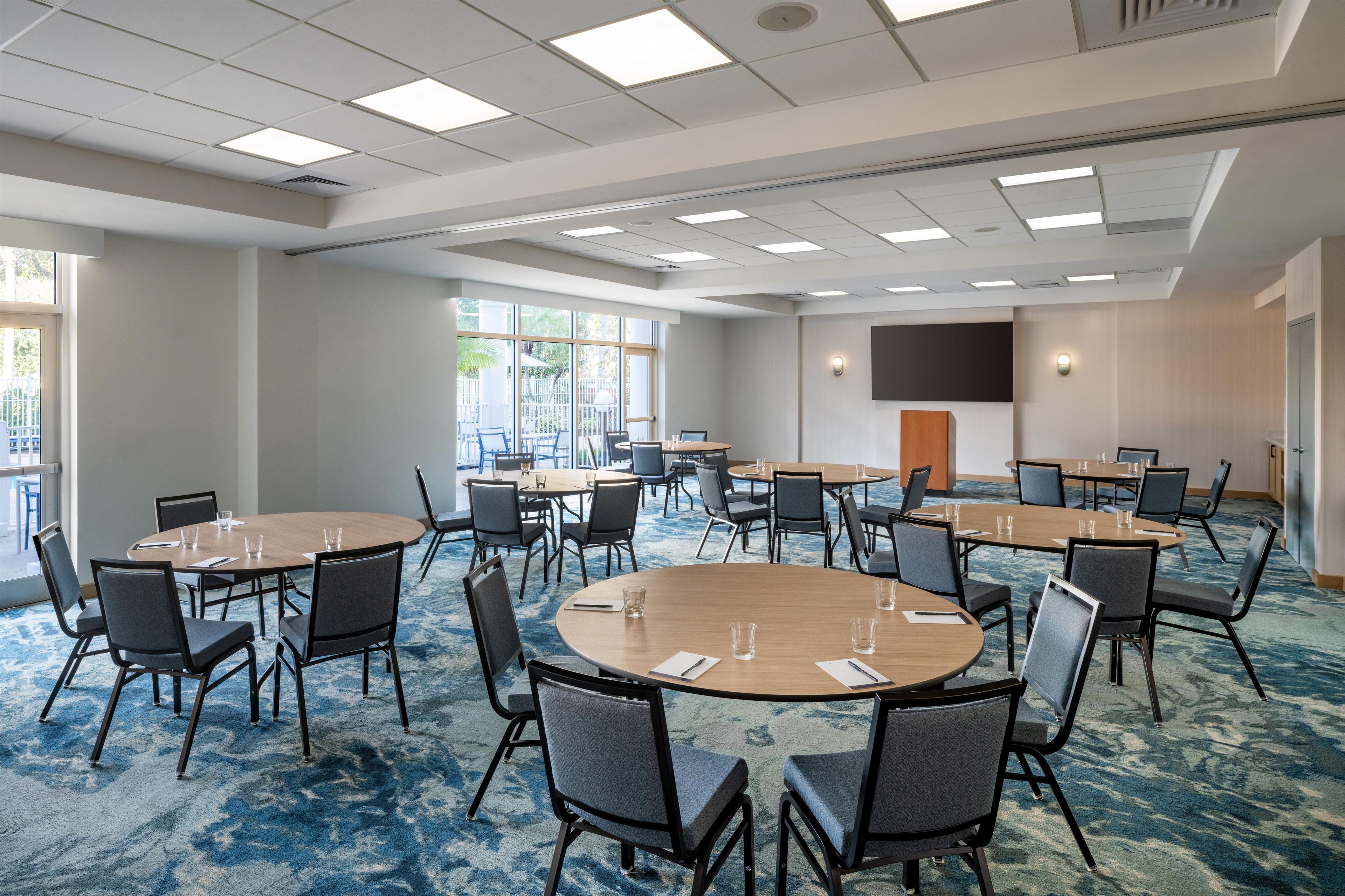 A conference room with multiple tables and chairs, large windows, and a flat-screen TV mounted on the wall.