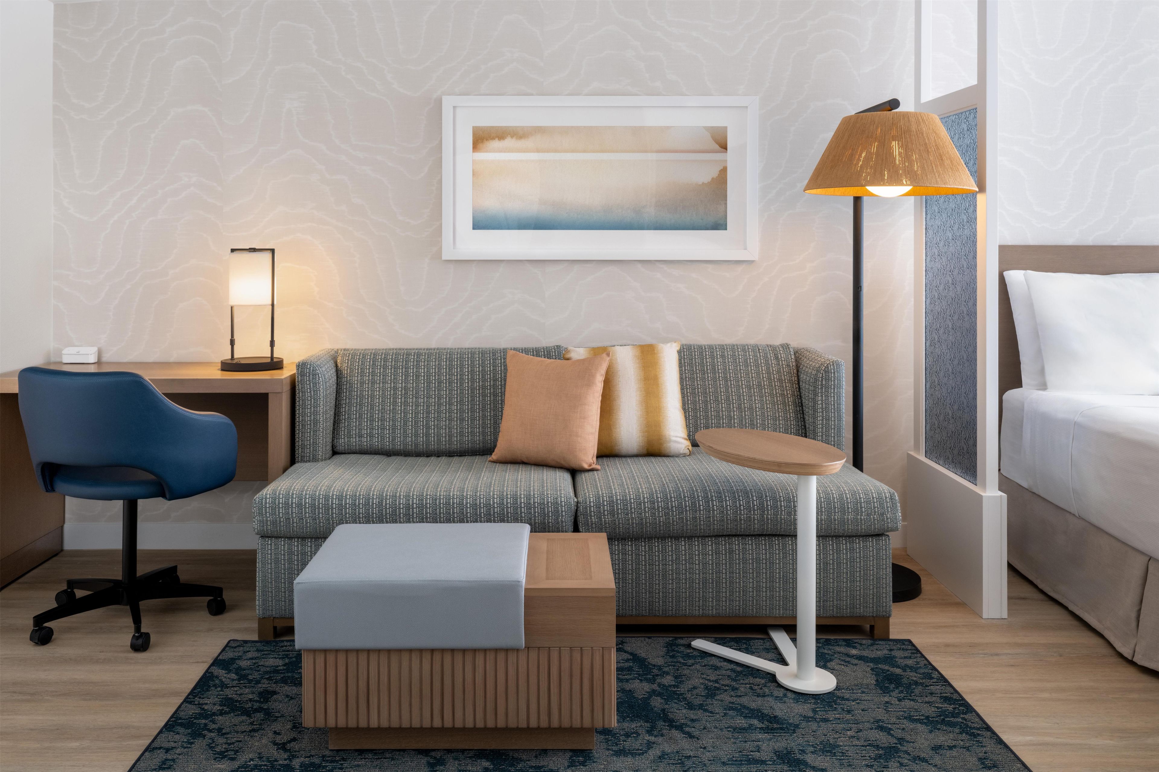 Una habitación de hotel moderna con un sofá gris estampado, una pequeña mesa auxiliar de madera, una silla de oficina azul, una lámpara de pie y una obra de arte enmarcada en la pared.
