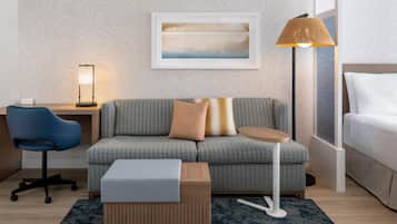 Una habitación de hotel moderna con un sofá gris estampado, una pequeña mesa auxiliar de madera, una silla de oficina azul, una lámpara de pie y una obra de arte enmarcada en la pared.