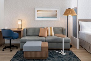 Una habitación de hotel moderna con un sofá gris estampado, una pequeña mesa auxiliar de madera, una silla de oficina azul, una lámpara de pie y una obra de arte enmarcada en la pared.