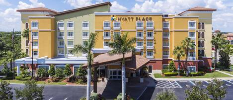 A multi-story hotel with a yellow facade, named "Hyatt Place." It features a central entrance with a covered walkway, palm trees, and a parking lot with marked spaces.