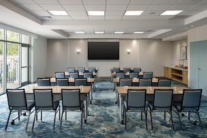 A conference room with a large screen, multiple tables, and chairs, equipped with overhead lighting.