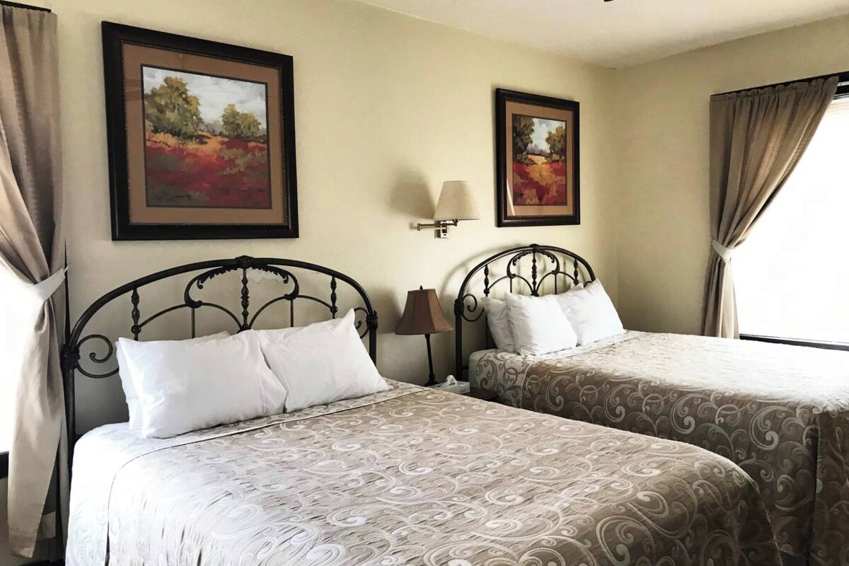 A hotel room with two beds, each with a patterned bedspread, a ceiling fan, and two framed paintings on the walls.