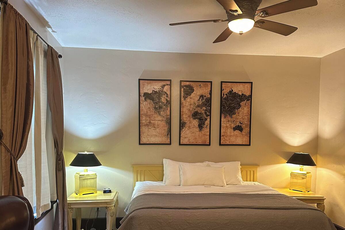 A hotel room with a bed, two bedside tables with lamps, a leather armchair, a red patterned carpet, and three world map wall art pieces.