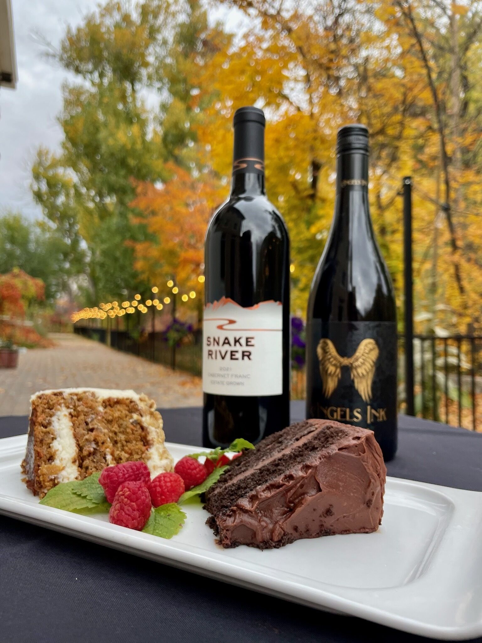 Une assiette avec une part de gâteau et des framboises, une bouteille de vin et une autre bouteille avec une étiquette.