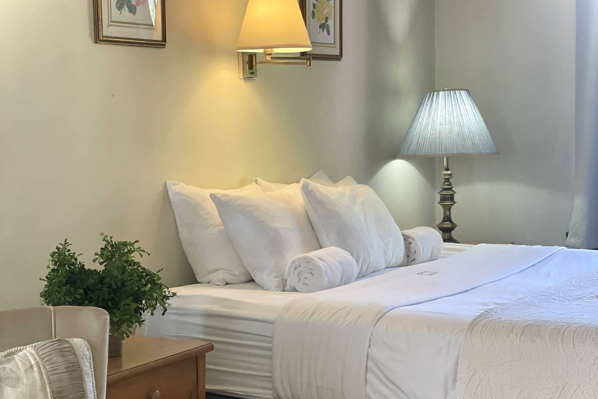 A neatly arranged hotel room with a bed, bedside lamps, a nightstand, a chair, and two framed pictures on the wall.