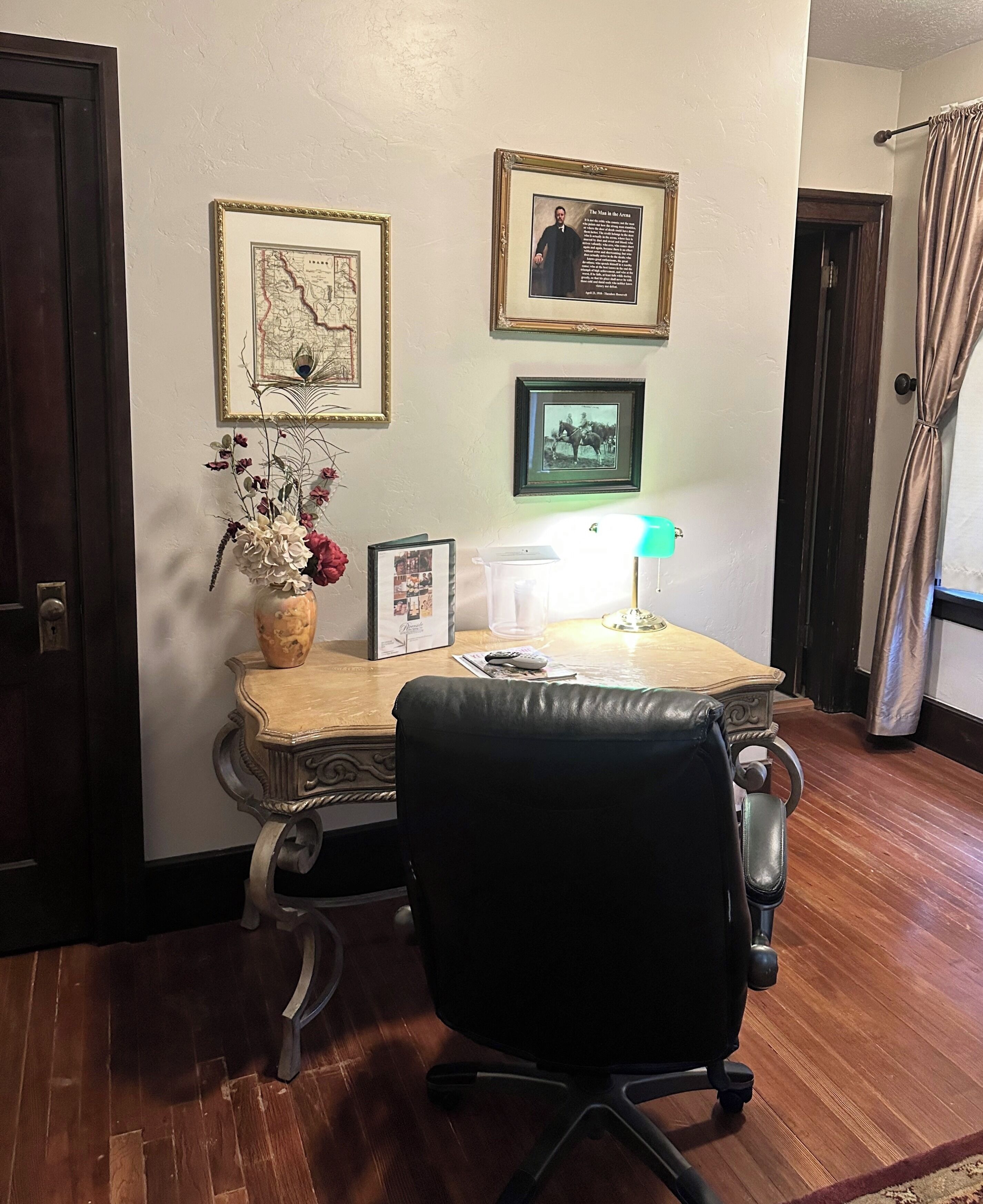 Una habitación con escritorio, silla, un jarrón con flores, cuadros en la pared y una lámpara.