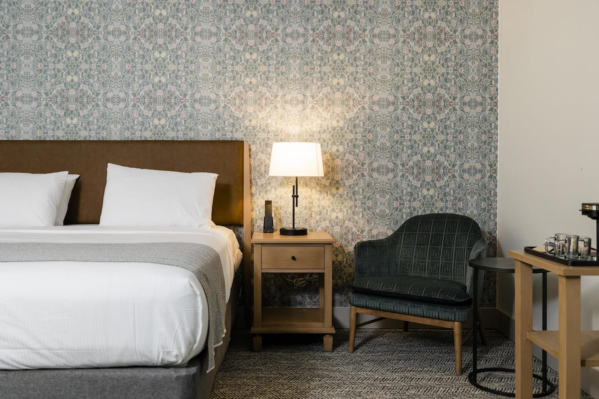 A hotel room with a bed, a nightstand with a lamp, a chair, and a small table.