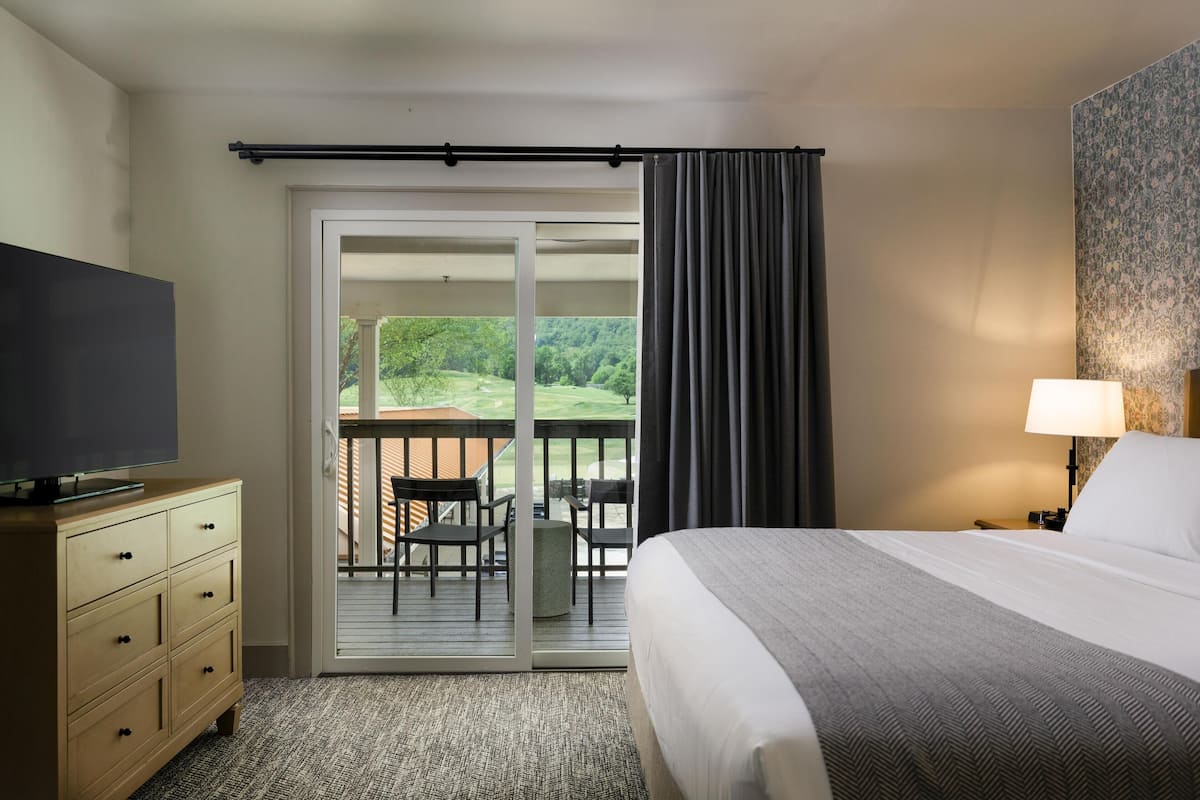 A hotel room with a bed, a TV on a dresser, a balcony with chairs, and a view of a green landscape.