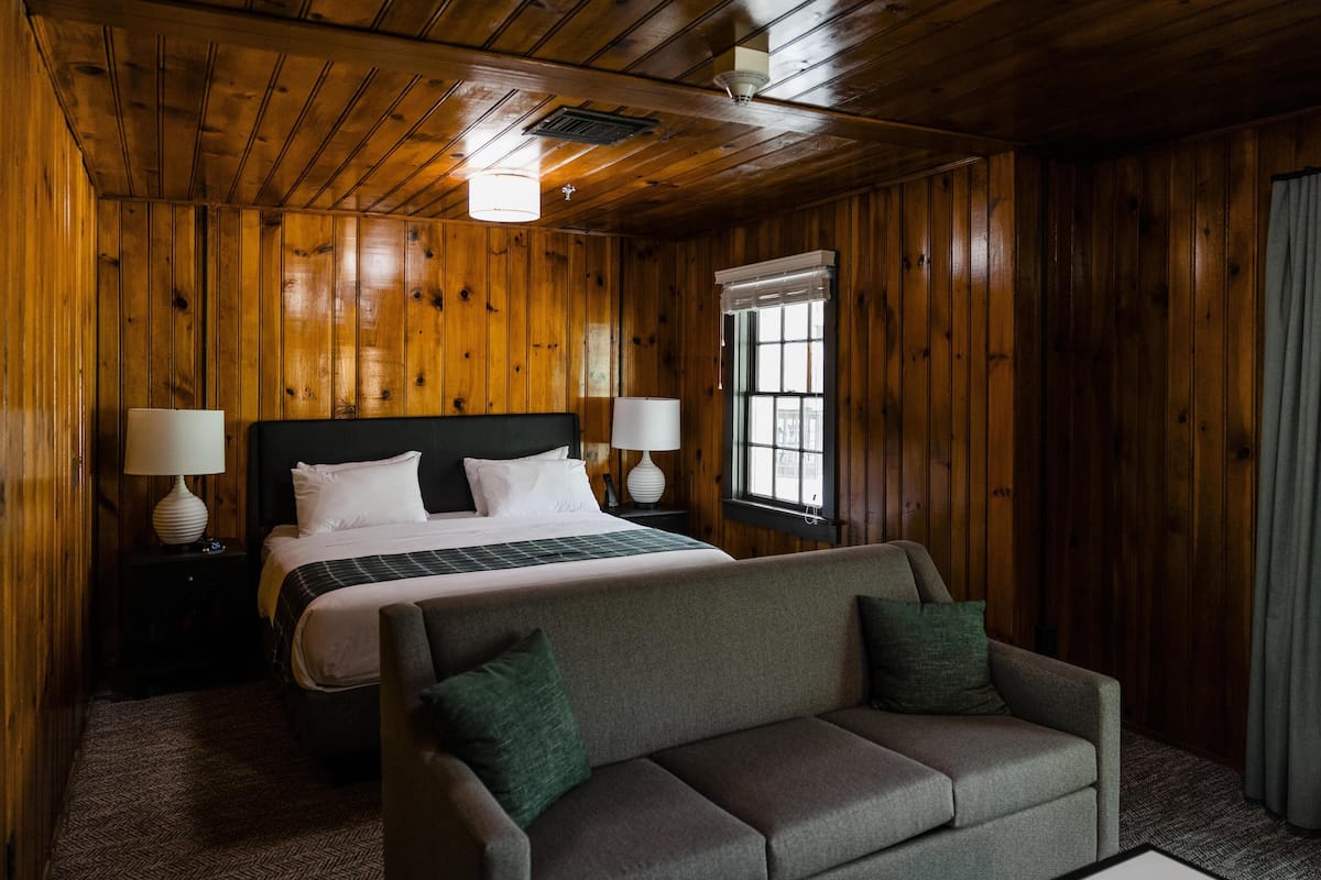 A wooden-paneled room with a bed, two bedside tables with lamps, a sofa, and a window with curtains.