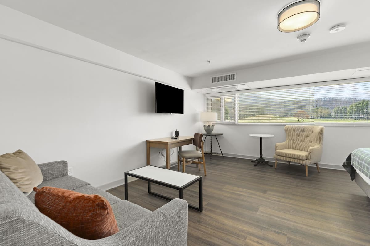 A modern hotel room with a grey sofa, a small table, a chair, a bed, a TV mounted on the wall, and a window with blinds.