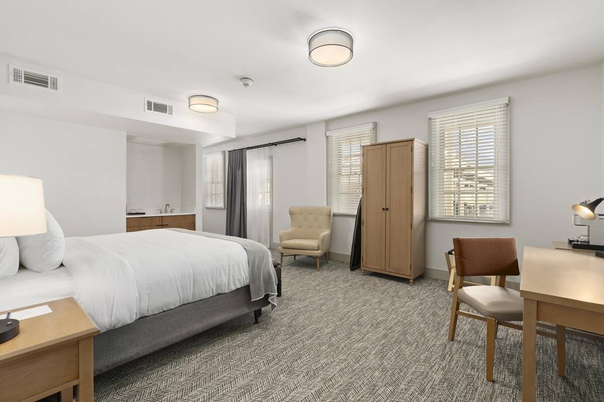 A hotel room with a bed, a desk, a chair, a wardrobe, and a window with blinds.