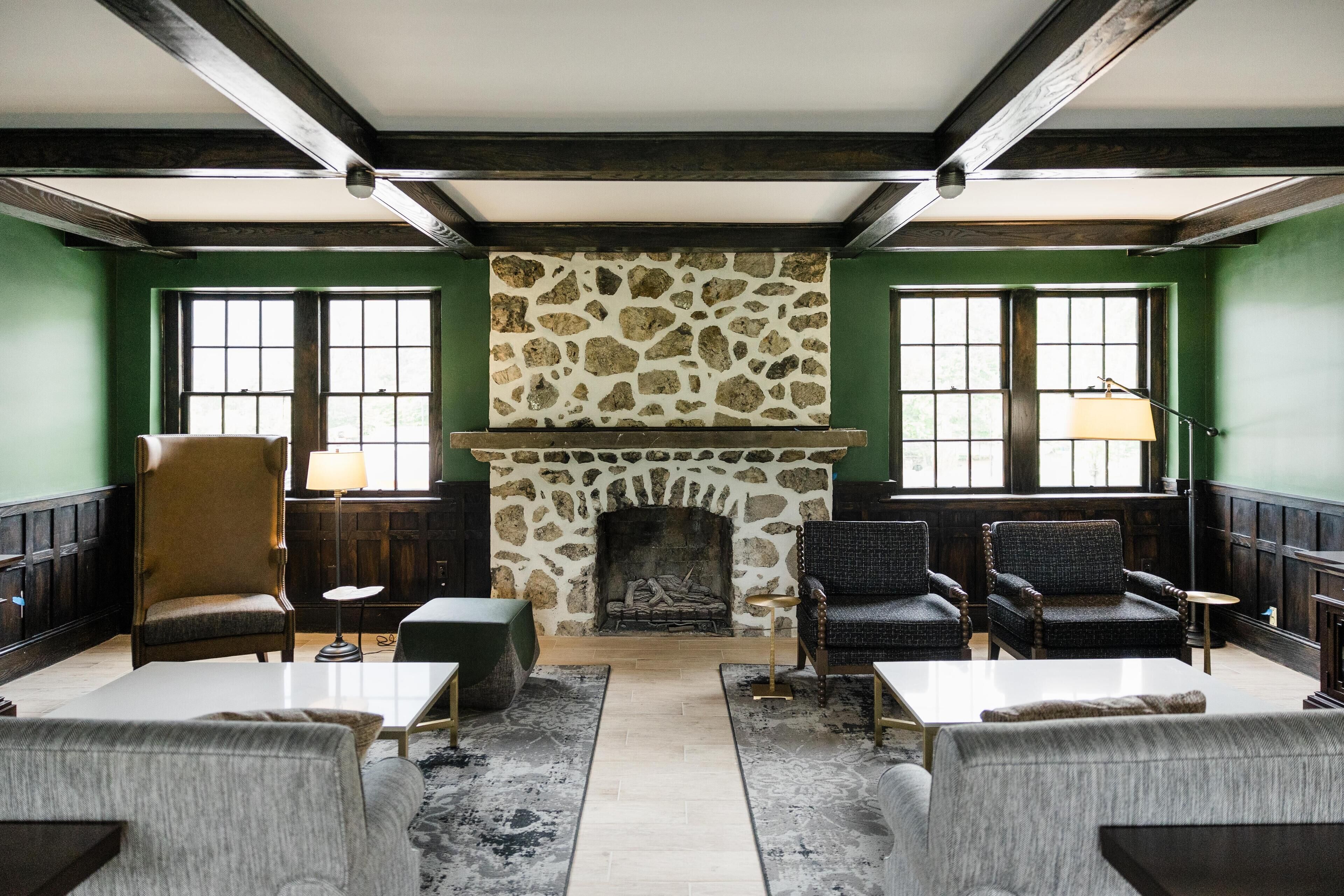 A room with a stone fireplace, green walls, and wooden beams. There are four chairs and two tables.