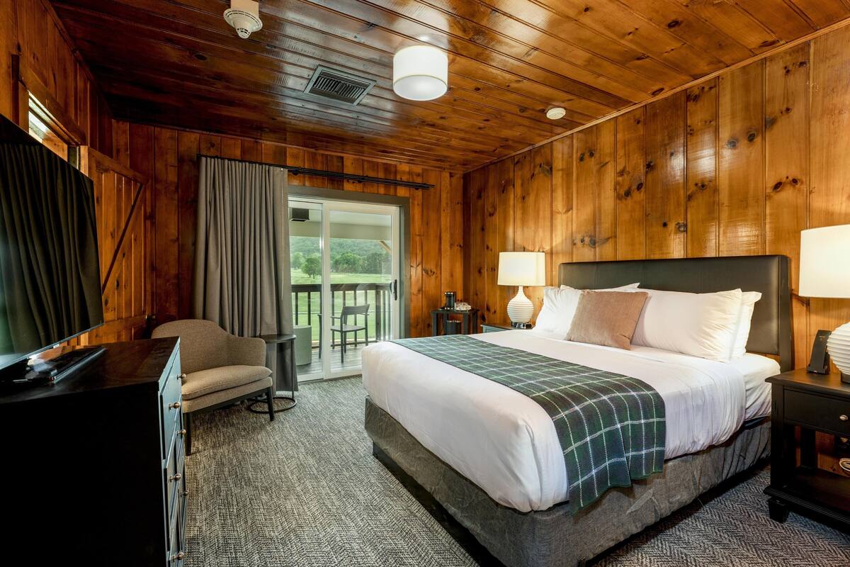 A wooden-paneled room with a bed, a desk, a chair, and a view through a glass door.
