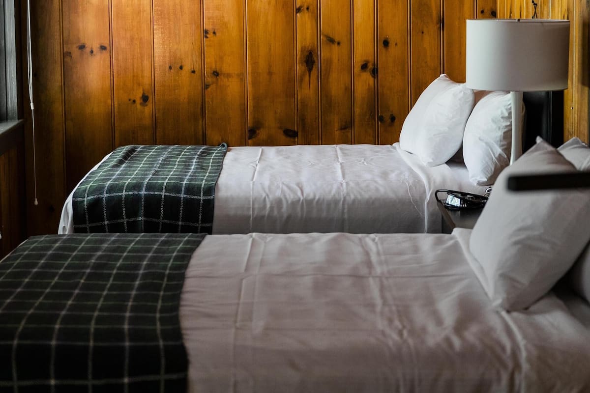 A room with wooden panel walls, two beds with white bedding, a nightstand with a lamp, and a window with curtains.