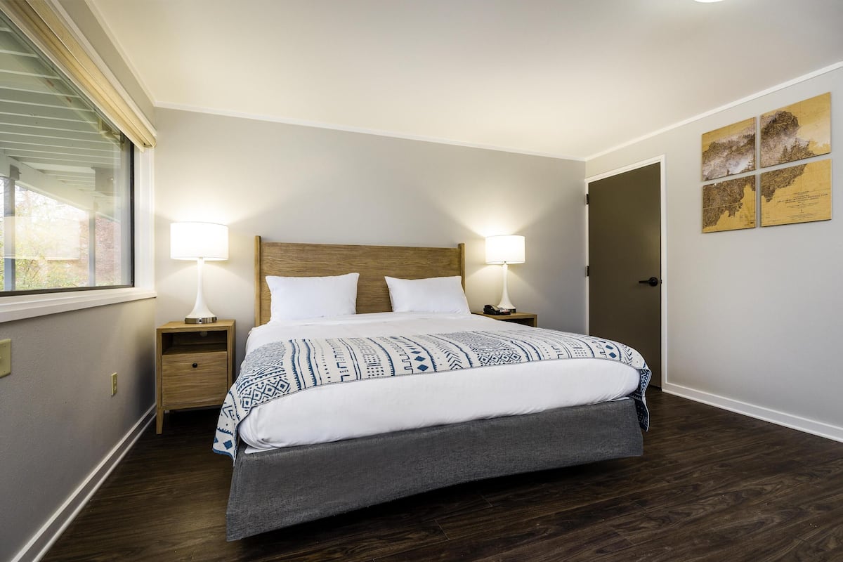 A bedroom with a large bed, two bedside tables with lamps, a window with blinds, and a door leading to another room.