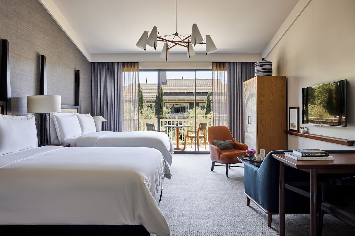 Habitación de hotel con dos camas, un escritorio con silla, televisión y balcón con mesa y sillas.