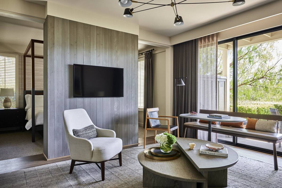 Una sala de estar moderna con un televisor de pantalla plana montado en una pared de paneles de madera, un área de estar acogedora con un banco y sillones, una mesa de centro con objetos decorativos, y ventanas amplias con vistas a la vegetación exterior.