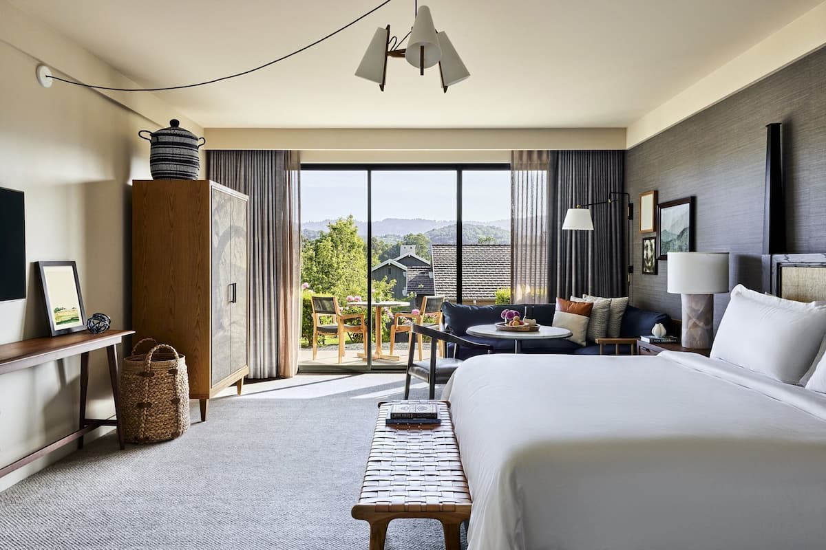 Habitación de hotel con cama, escritorio, silla, mesita, lámpara y vistas al exterior.