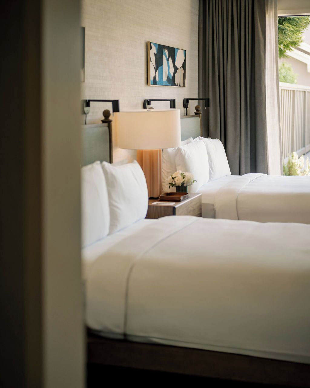 Habitación de hotel con dos camas, una mesita de noche, una lámpara, un cuadro en la pared y vistas al exterior.