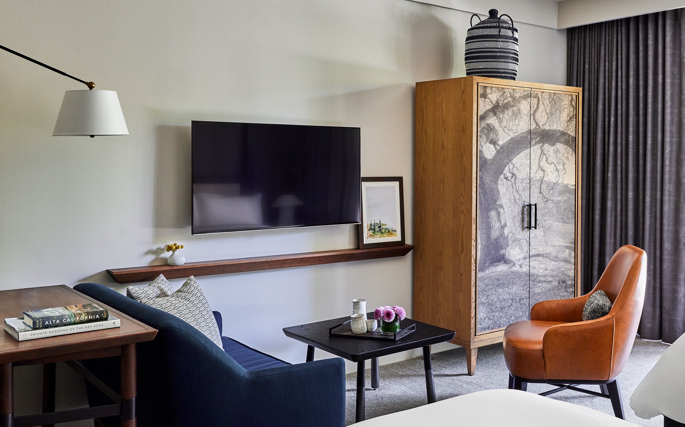 A modern hotel room with a flat-screen TV, a wooden cabinet with a nature-themed door, a sofa, a chair, and a small table with a tray.