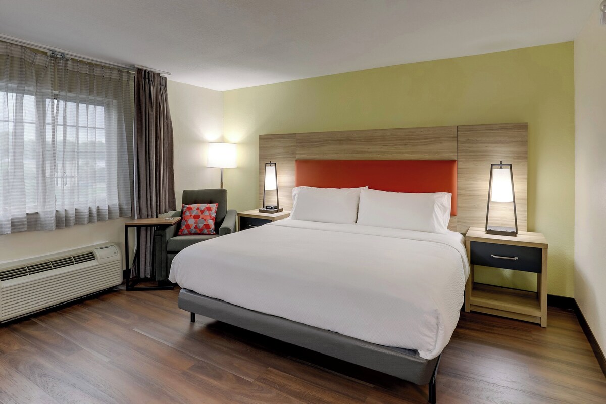 A hotel room with a large bed, two bedside tables with lamps, a chair, a heater, and a window with curtains.