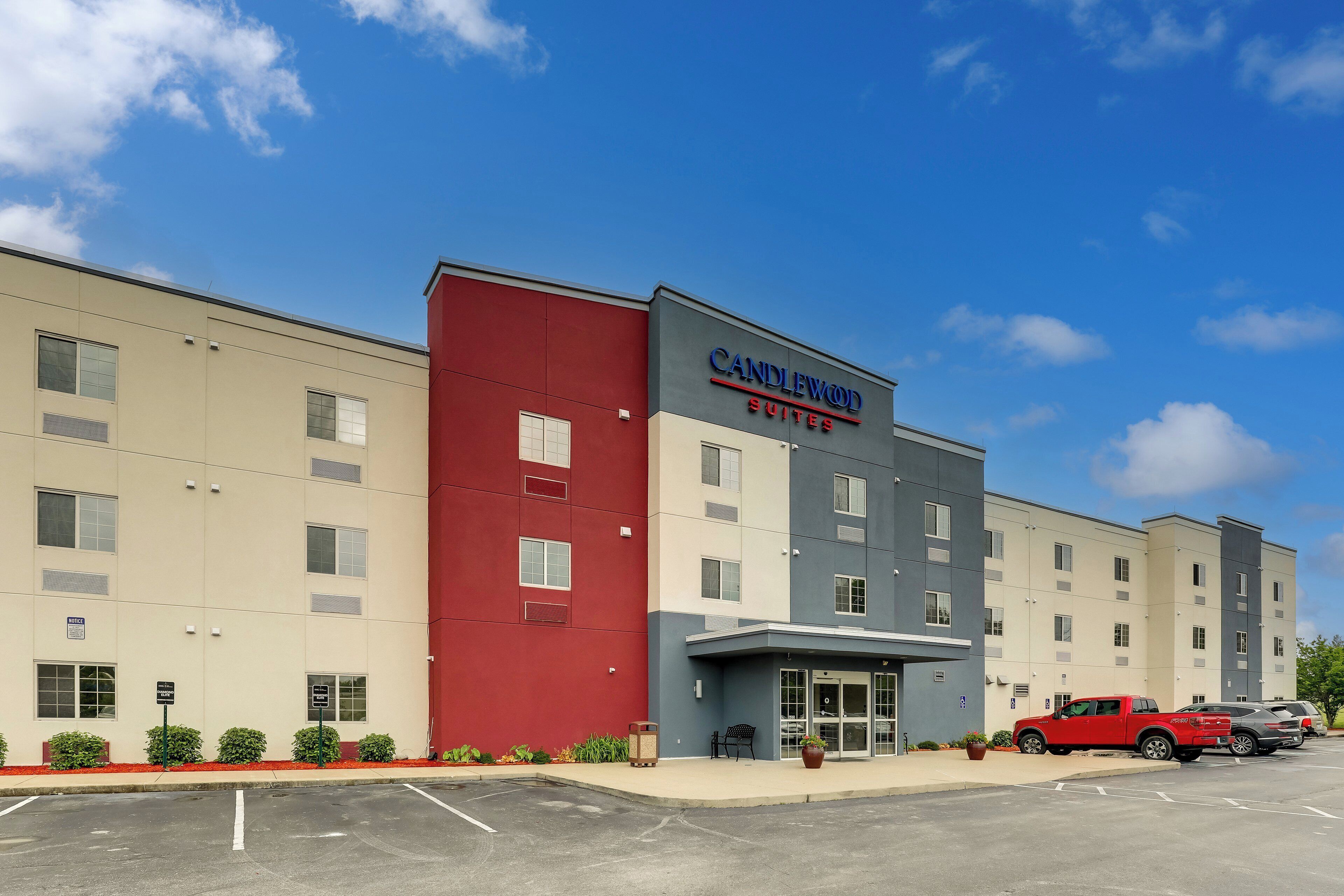 A multi-story hotel with a red and grey facade, a parking lot with several cars, and a clear blue sky.