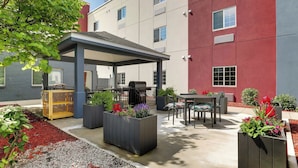 A covered outdoor seating area with tables and chairs, surrounded by planters with flowers, and a building with multiple windows and a red facade. - Candlewood Suites Lexington by IHG (Lexington)