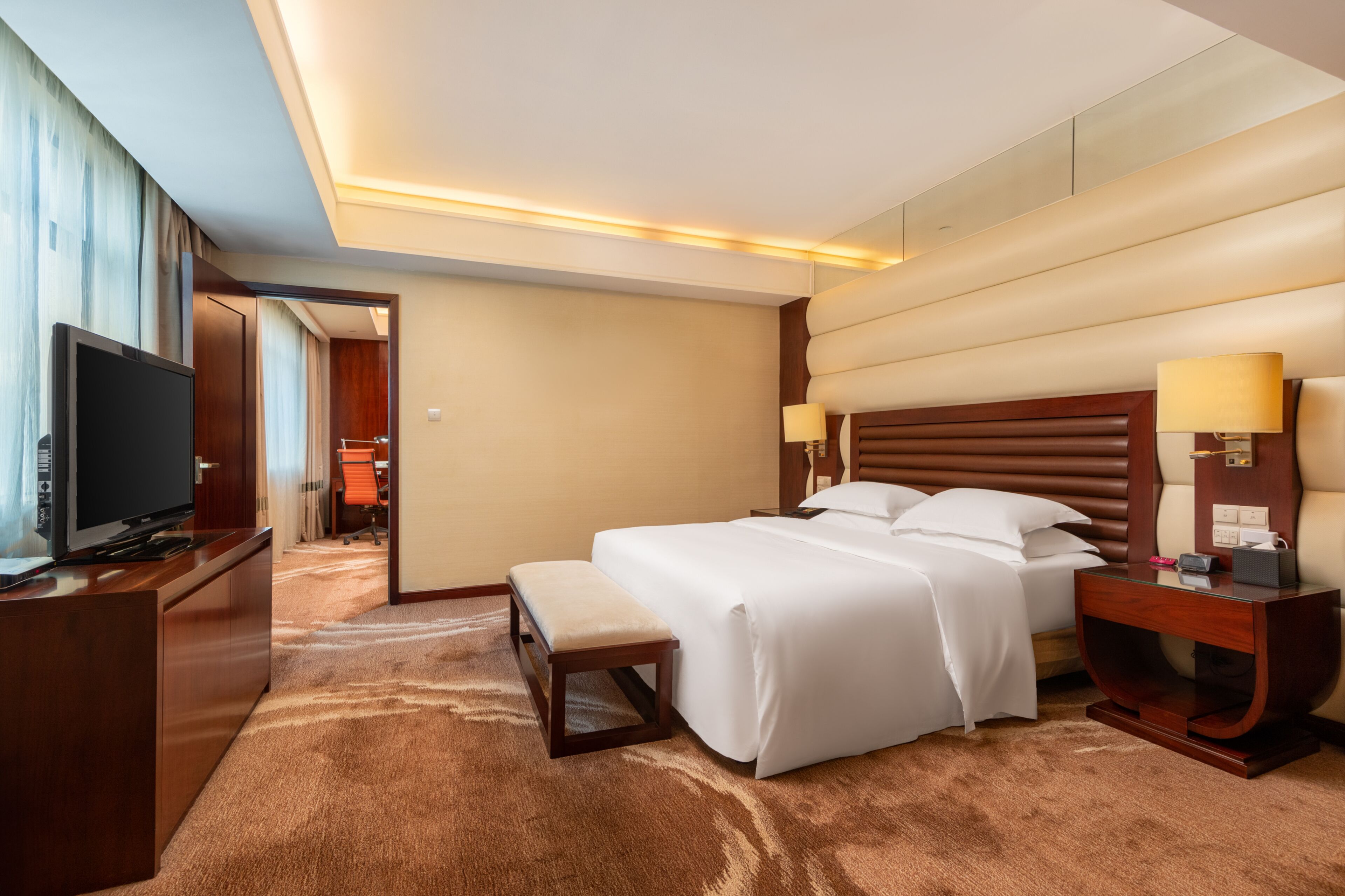 Una habitación de hotel con una cama grande, lámparas de noche, un televisor, un escritorio y un banco.