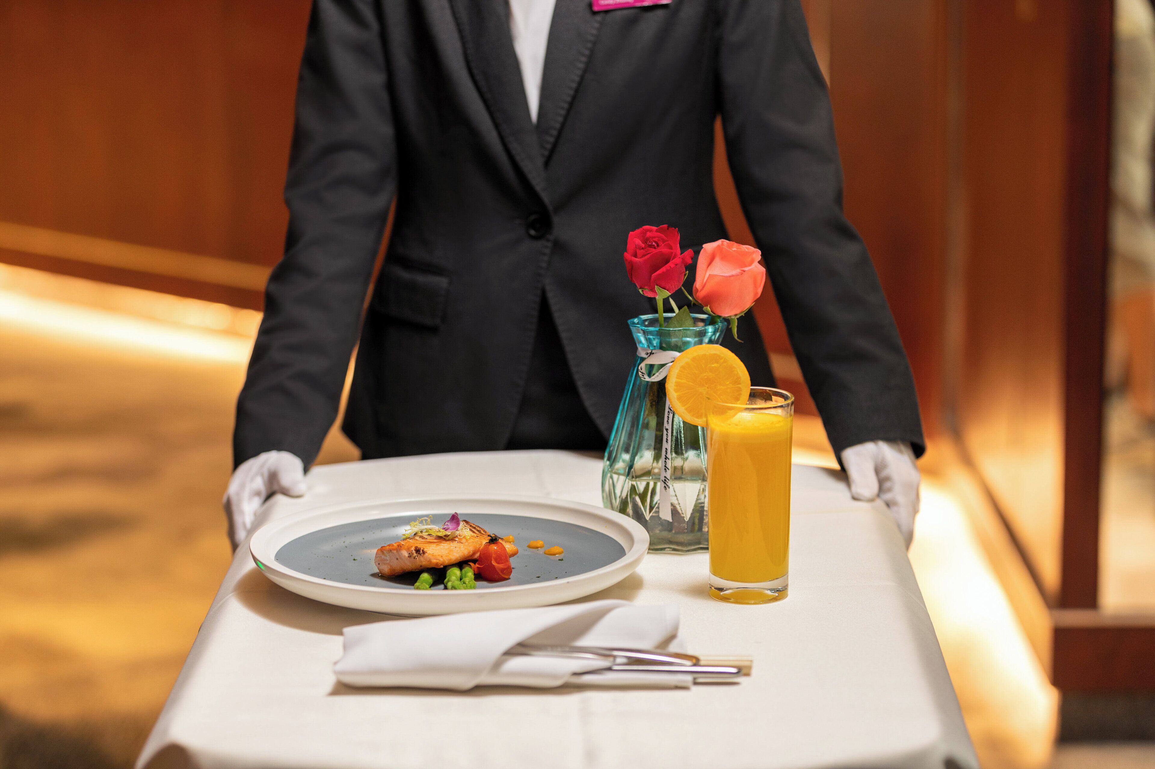 Un serveur en costume noir apporte une assiette de nourriture et un verre de jus d’orange à une table.