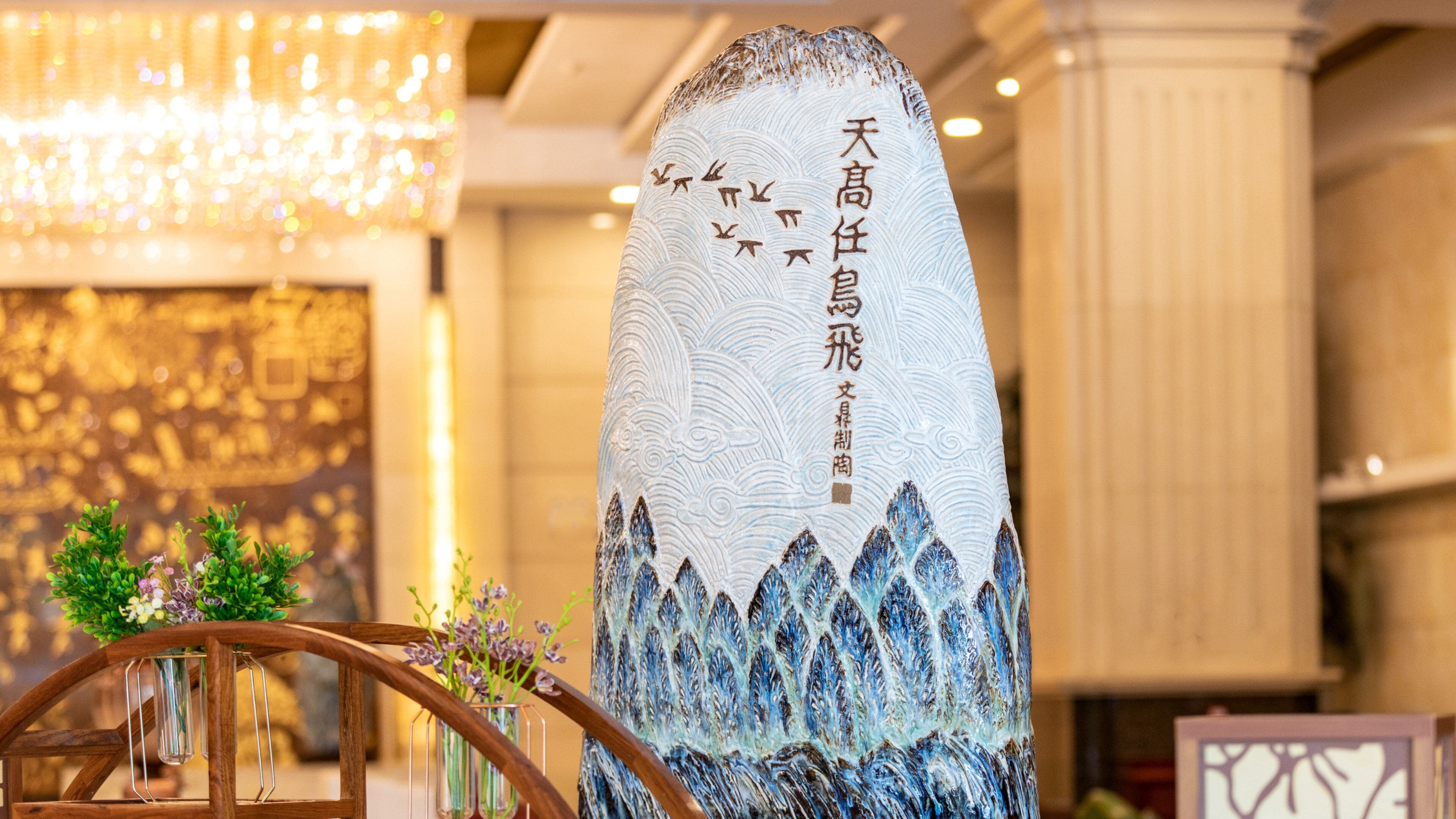 A tall, intricately designed pillar with Chinese calligraphy, set in a well-lit indoor space with a chandelier and a wooden chair with a vase of flowers.