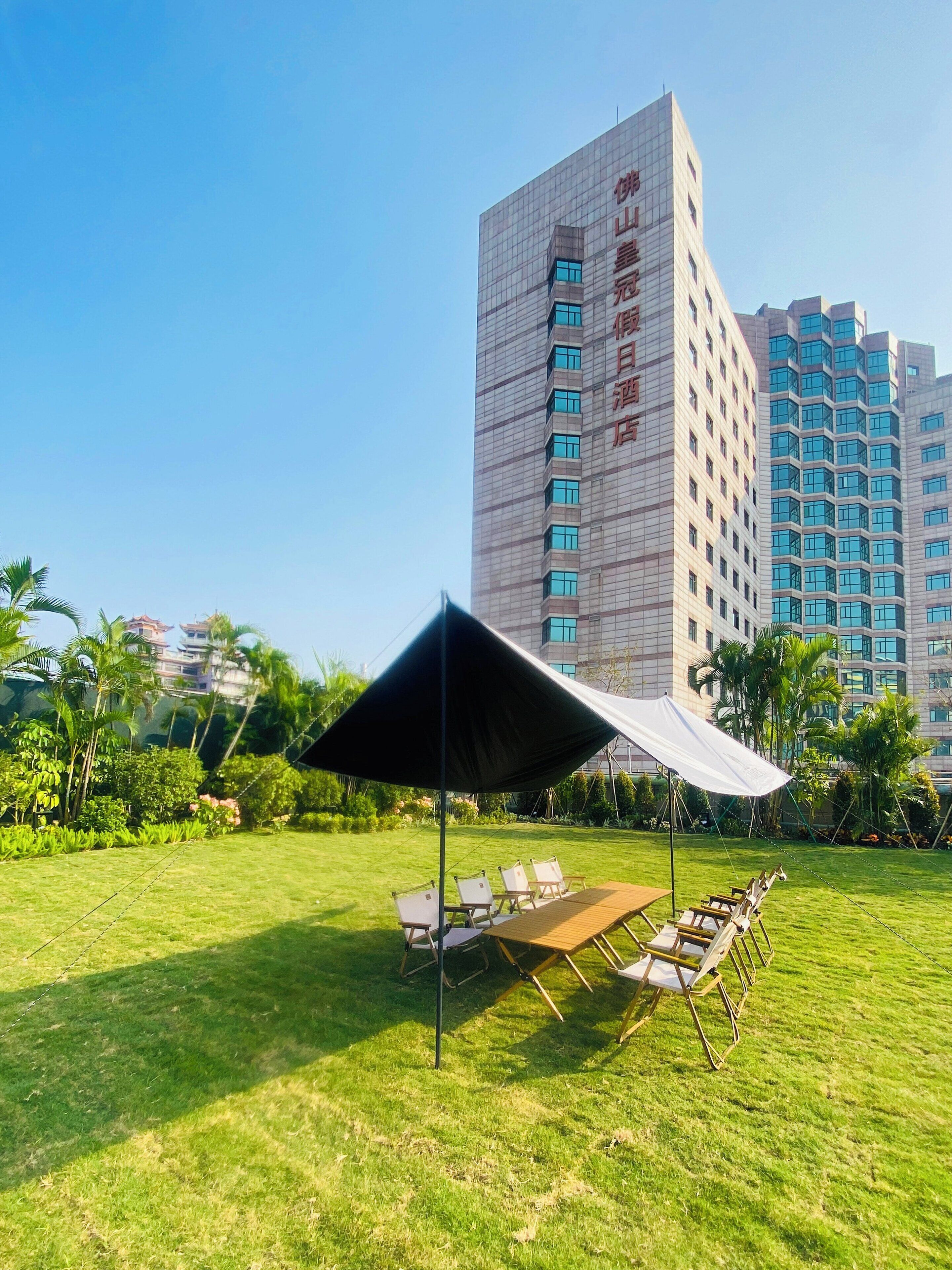 A grassy outdoor area with a tent and chairs, surrounded by modern buildings with visible signage.