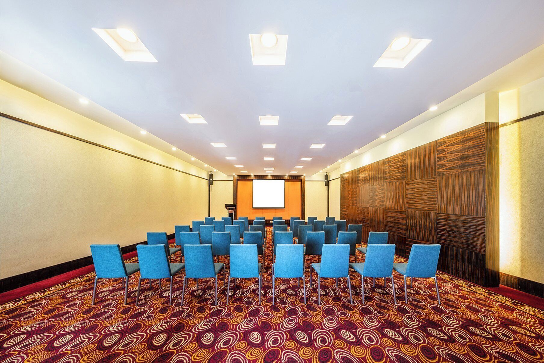 A conference room with rows of blue chairs, a patterned carpet, and a projector screen at the front.