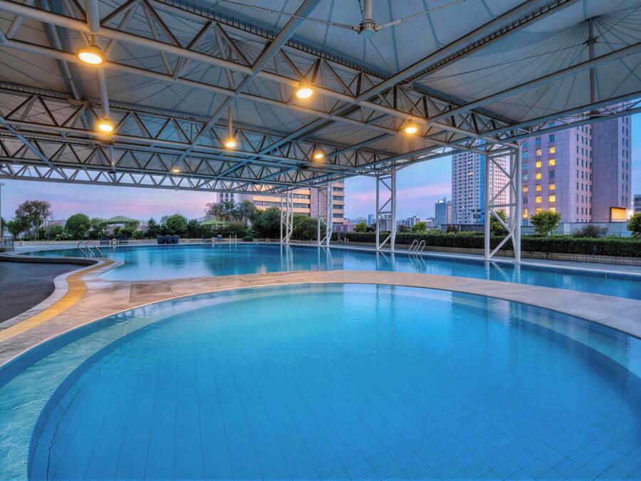 Una gran piscina cubierta con techo, rodeada de jardines con árboles y un paisaje urbano al fondo.