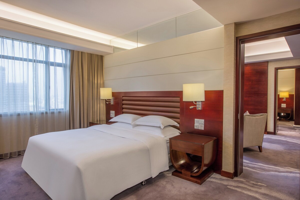 A hotel room with a large bed, two bedside lamps, a wooden headboard, and a view of the city through large windows.