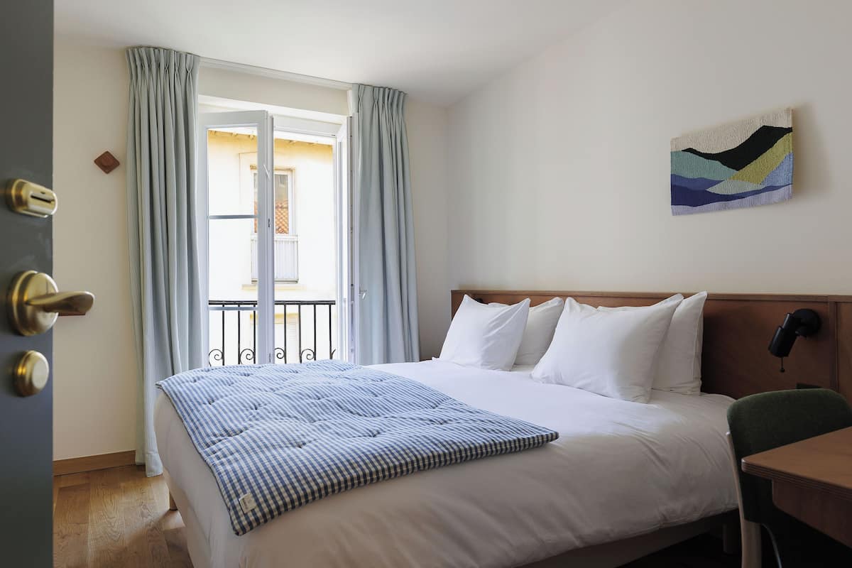 A hotel room with a bed, a desk, a chair, a painting on the wall, and a view of the outside through the window.