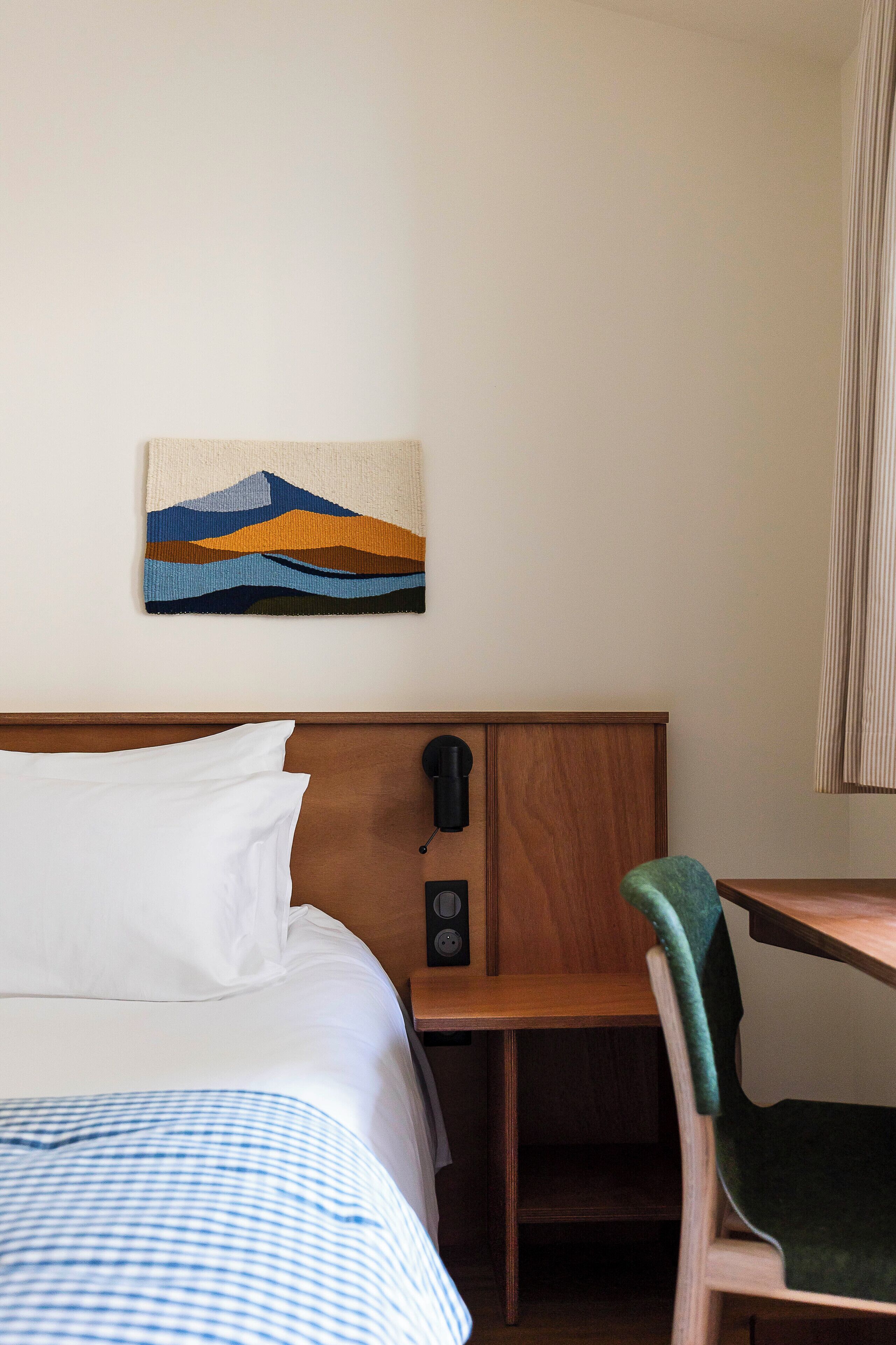 A hotel room with a bed, a desk, a chair, a wall-mounted phone, and a painting.