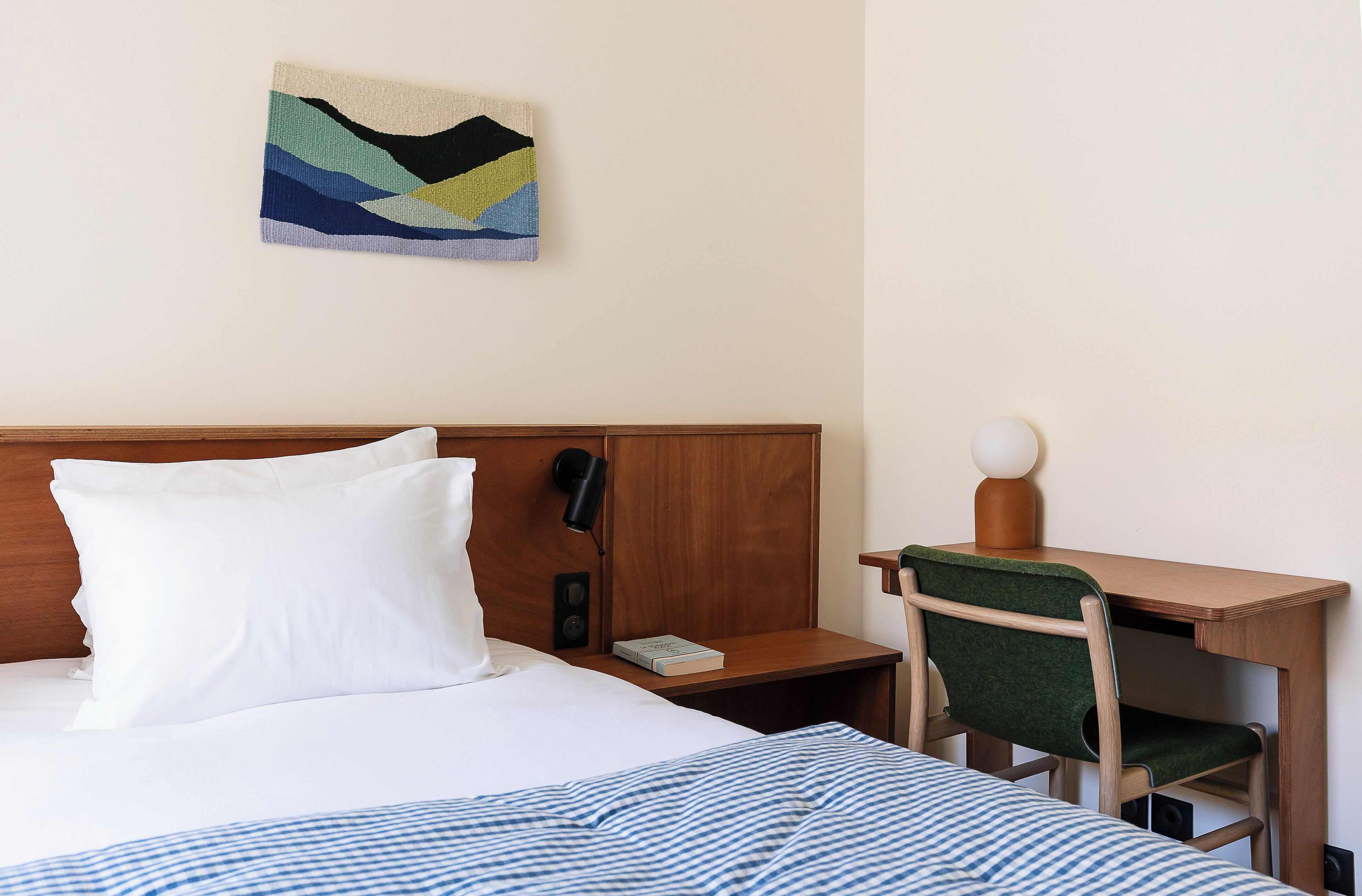 A neatly made bed with white bedding, a wooden headboard, a bedside table with a lamp, a chair, and a small desk with a lamp.