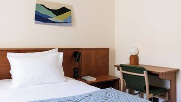 A neatly made bed with white bedding, a wooden headboard, a bedside table with a lamp, a chair, and a small desk with a lamp.