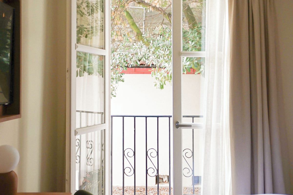A bedroom with a bed, a chair, a desk, and an open door leading to a balcony with plants.