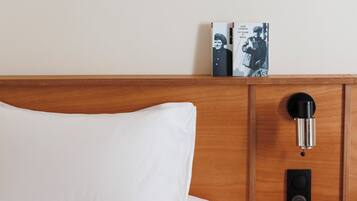 Habitación de hotel con cabecera de madera, almohada blanca, mesita de noche con lámpara y un libro en la pared.