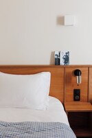Habitación de hotel con cabecera de madera, almohada blanca, mesita de noche con lámpara y un libro en la pared.