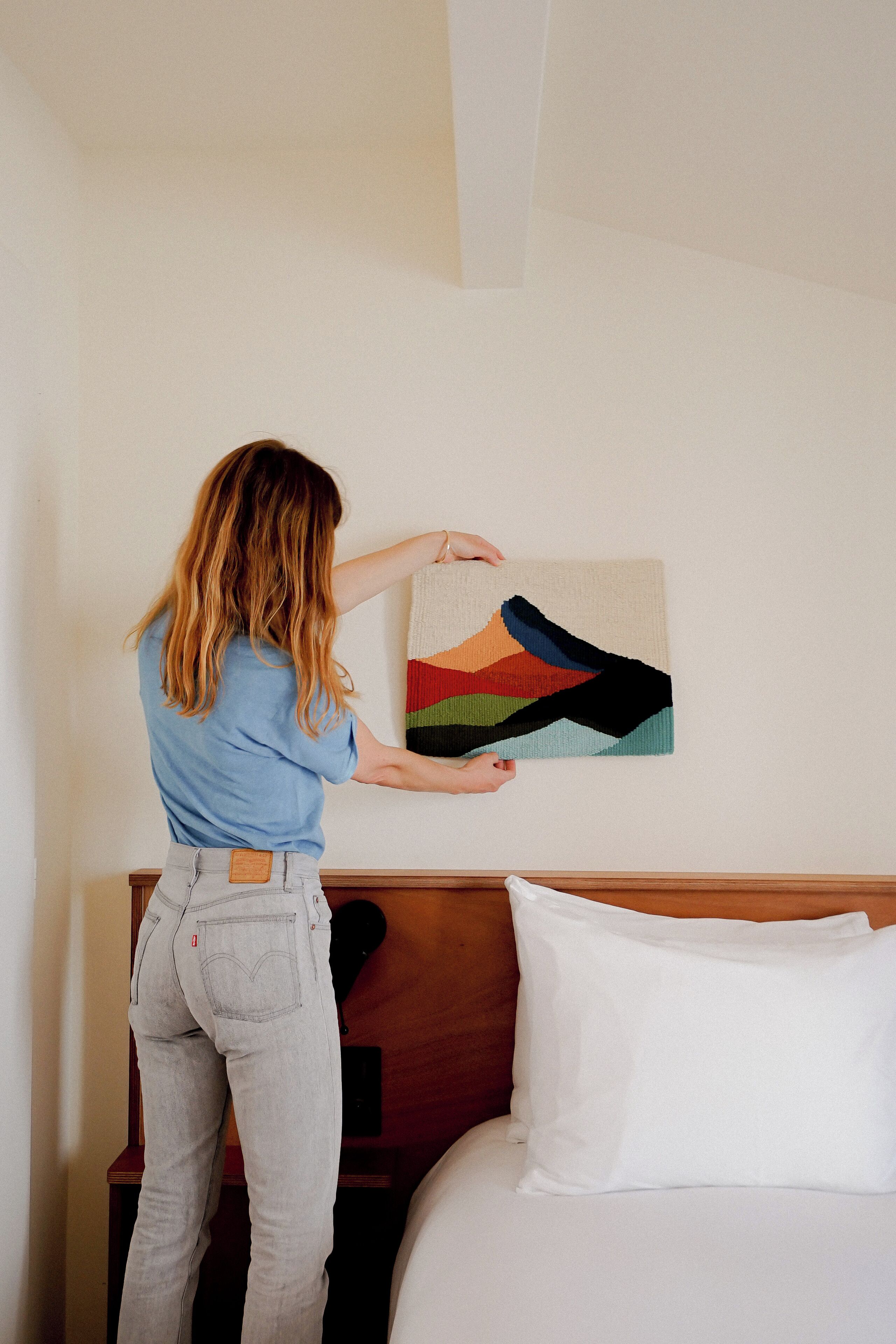 A person hanging a colorful abstract painting on a wall in a bedroom with a wooden headboard and white bedding.