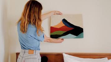 A person hanging a colorful abstract painting on a wall in a bedroom with a wooden headboard and white bedding.