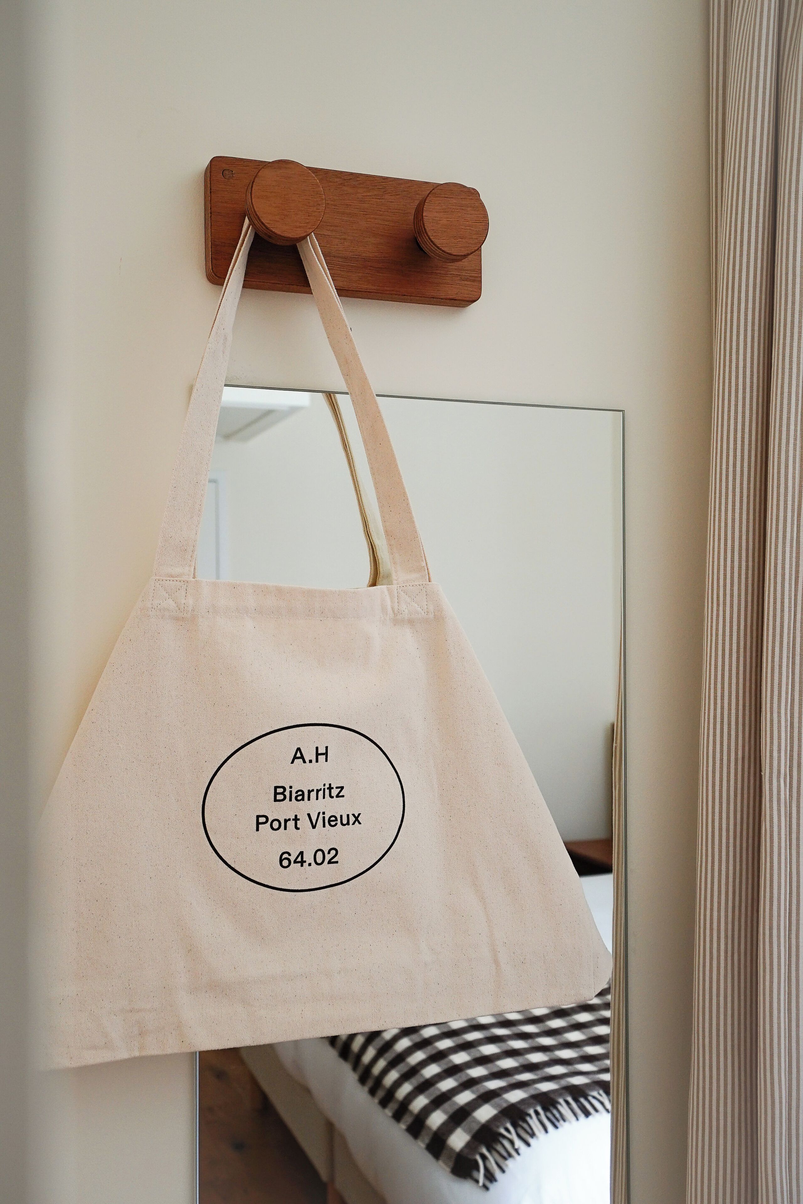Una bolsa de lona color beige colgada de un gancho de madera, con un espejo que refleja una cama y cortinas a rayas.