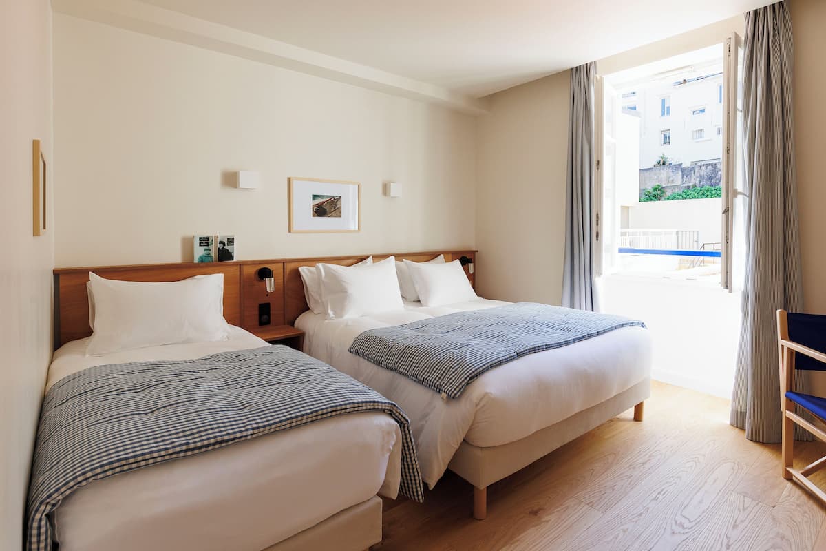 A hotel room with two beds, a wooden headboard, a small bedside table, a framed picture on the wall, and a view of the outside balcony.