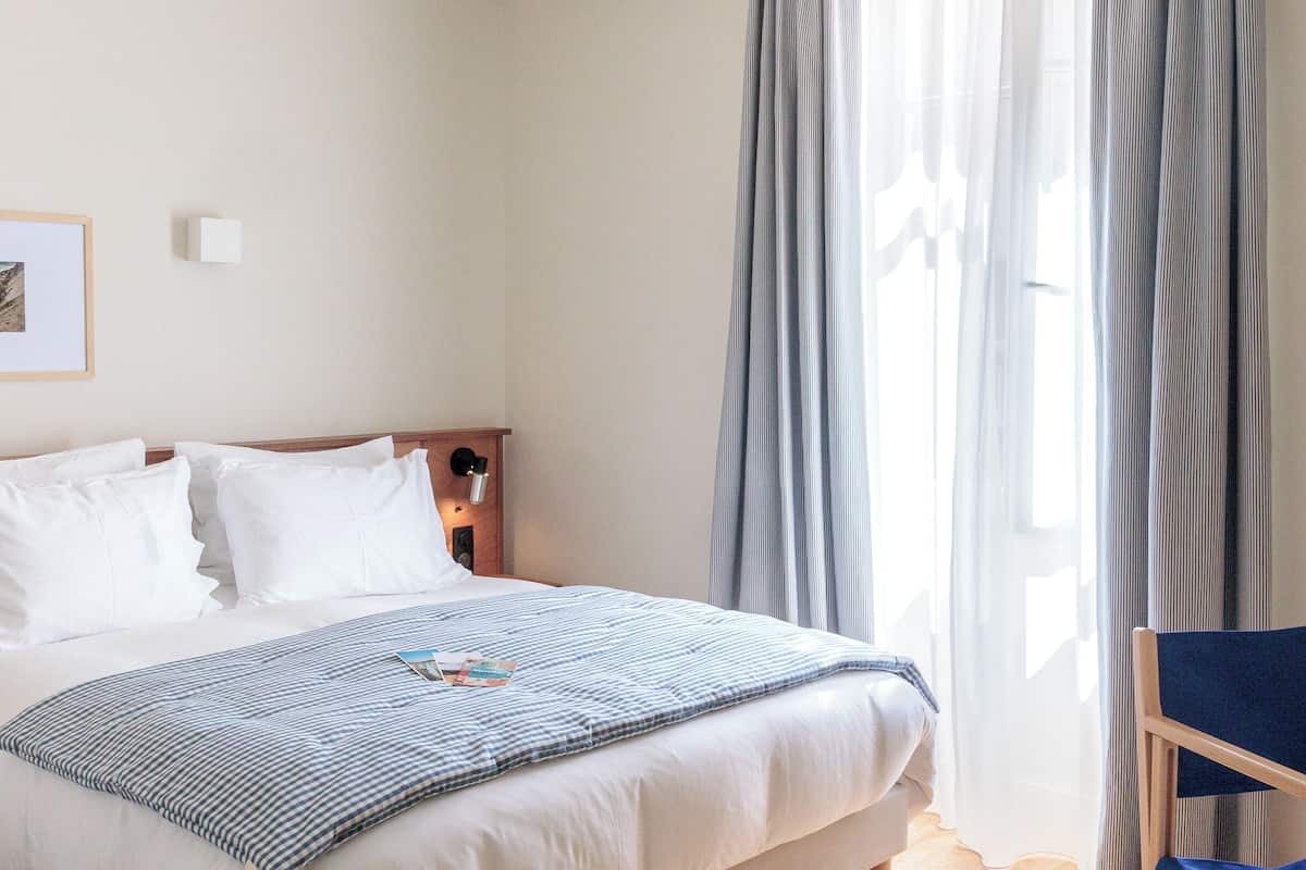 A hotel room with a bed, pillows, a bedside table with a lamp, a chair, and a window with curtains.