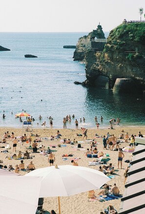 A crowded beach with people sunbathing and swimming, a rocky cliff with a statue, and a bridge connecting the land to the cliff. - Alfred Hotels Port-Vieux  (Biarritz)
